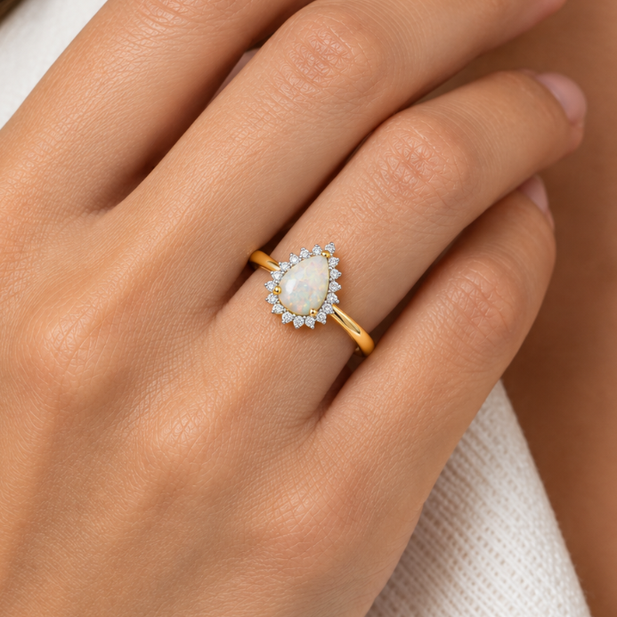 A close-up of a hand wearing a gold ring with a pear-shaped white stone surrounded by small clear stones, set against a soft white garment background.